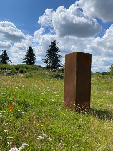 Säule auf einer Wiese Ein rostiger, aufrechter Block steht in einer blühenden Wiese unter bewölktem Himmel.