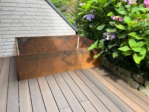 Hochbeet Eckiger Pflanzkasten aus Cortenstahl auf einer Holzterrasse, umgeben von Pflanzen.