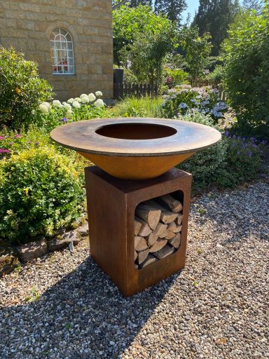 Plancha Grill 1000 mm Moderne Feuerschale aus Metall auf quadratischem Holzständer im Garten.