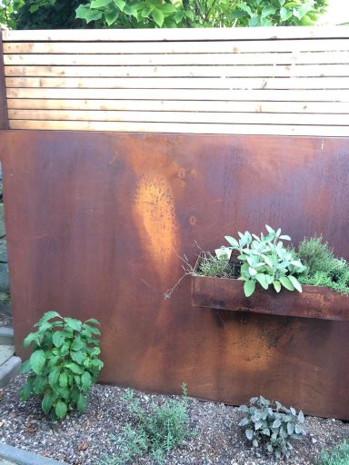 Sichtschutzelement Rostige Wand mit bepflanztem Blumenkasten und grünen Pflanzen im Vordergrund.